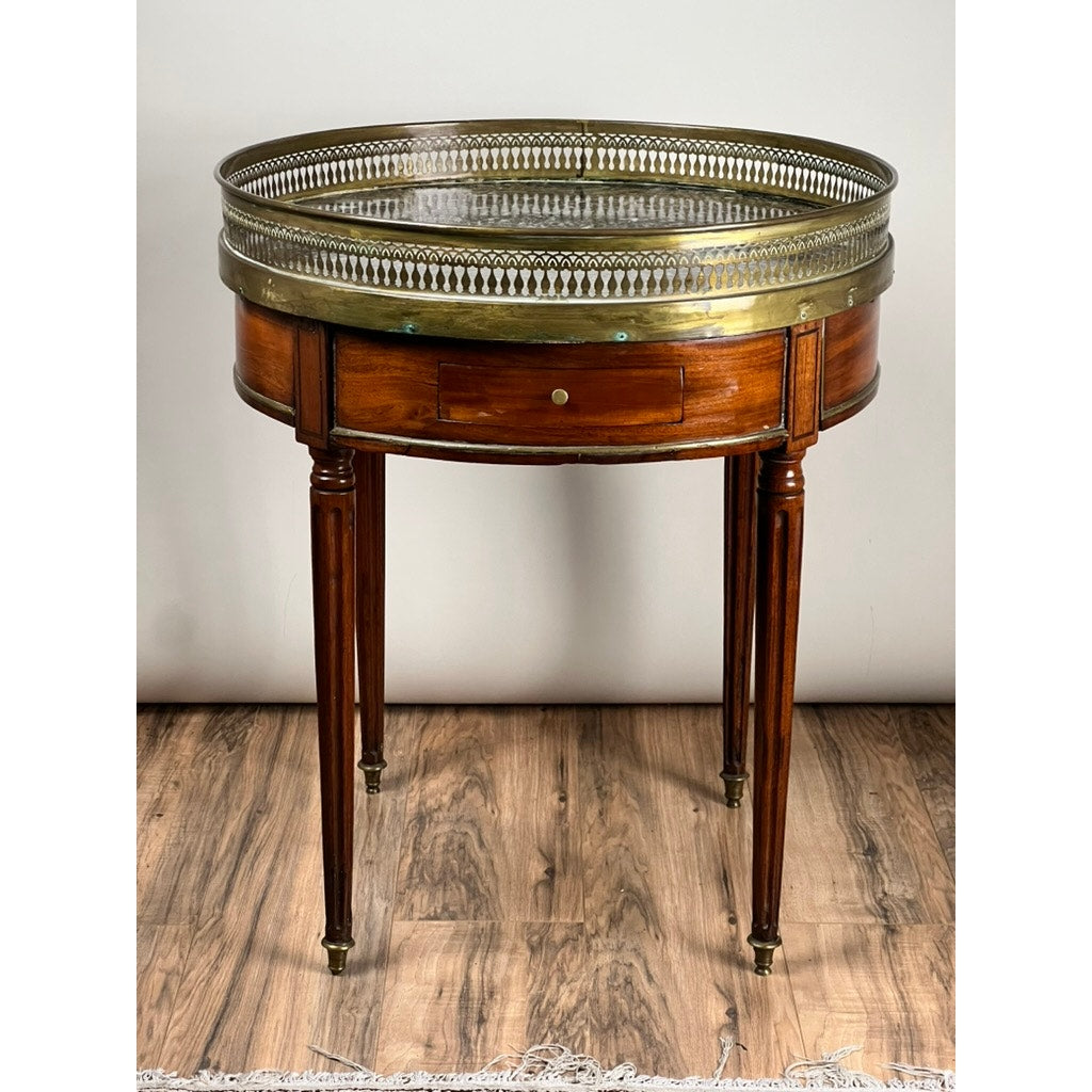 An Antique 19th Century French Louis XVI-Style Mahogany Bouillotte Table with a marble top and pierced brass gallery stands on a wooden floor against a plain light-colored wall.