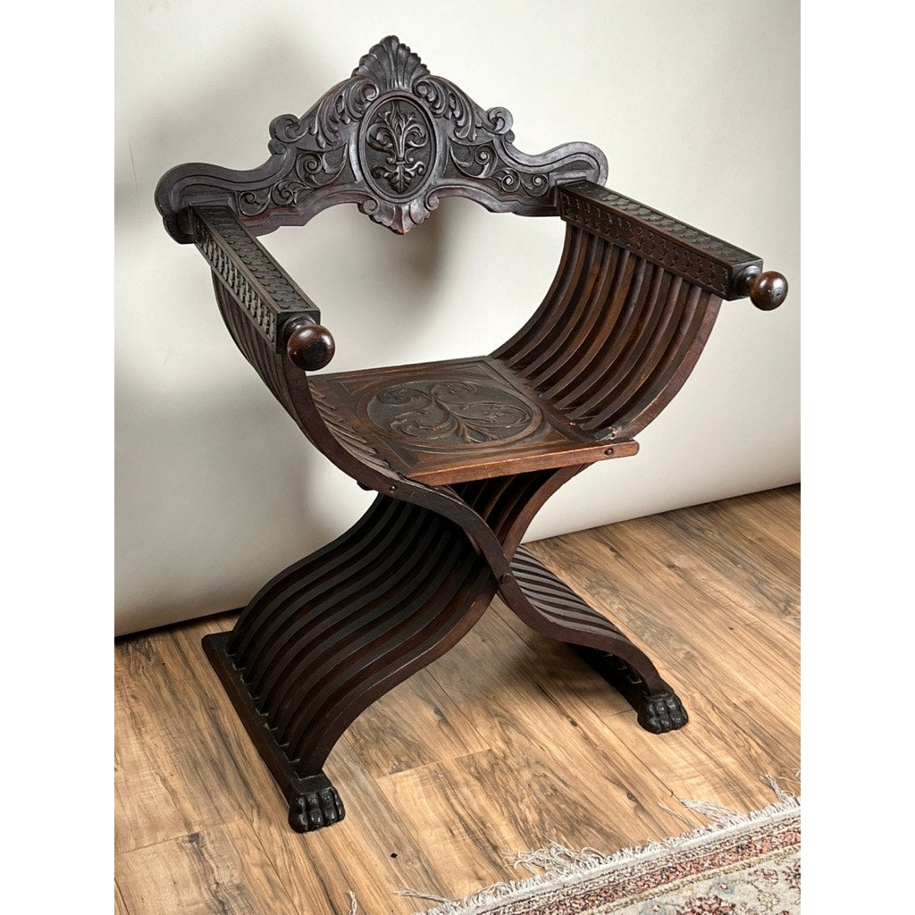 Antique Italian Renaissance Walnut Savonarola Chair (late 19th century) with a curved X-shaped frame, carved floral backrest and seat, and lion paw feet, displayed on a wooden floor near a rug. Brand: None/Unknown.
