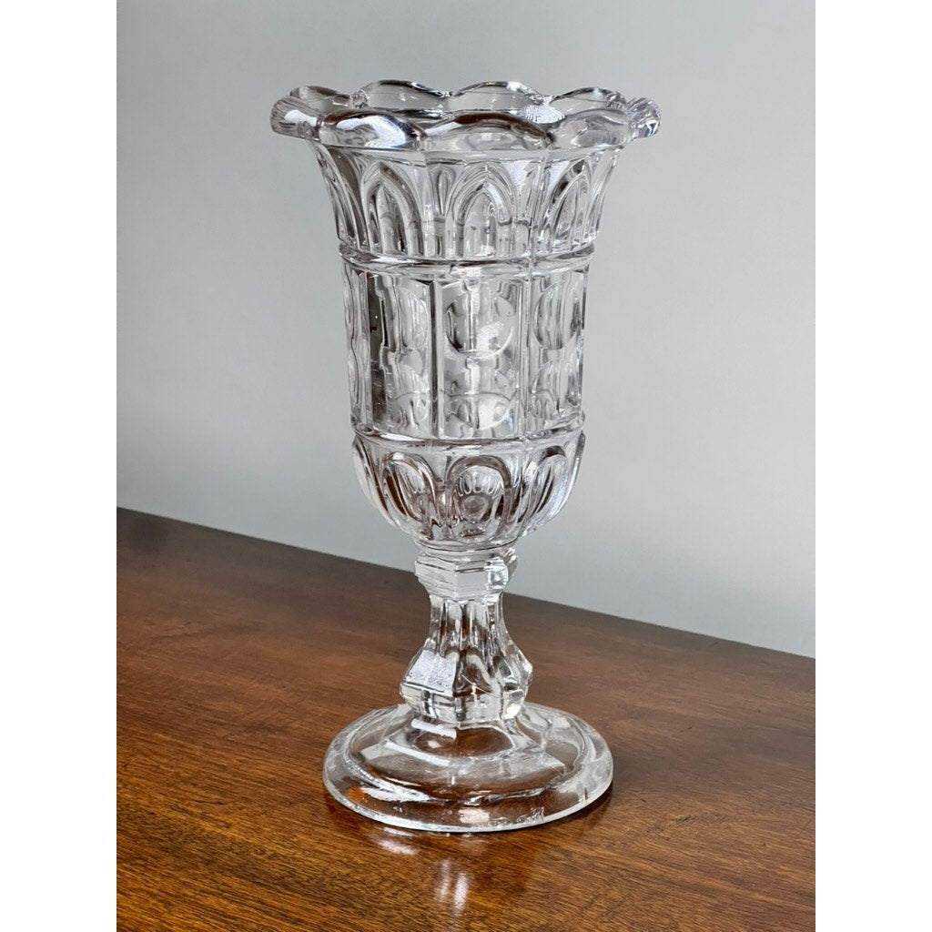 An Antique Boston & Sandwich Glass Co. Gothic Arch Pressed Glass Celery Vase (c. 1860–1880) by Sandwich Glass stands on a wooden surface against a plain light gray background, featuring an ornate fluted rim and decorative pattern.