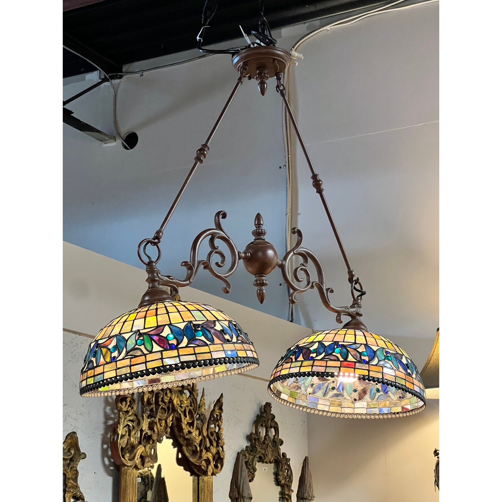 The Vintage Tiffany Style Leaded Stained Glass Double Pendant Billiard Island Light, featuring floral-patterned stained glass shades suspended from a brown ornate metal frame, hangs elegantly in a room with mirrors and white walls—making it an ideal choice for illuminating a kitchen island.