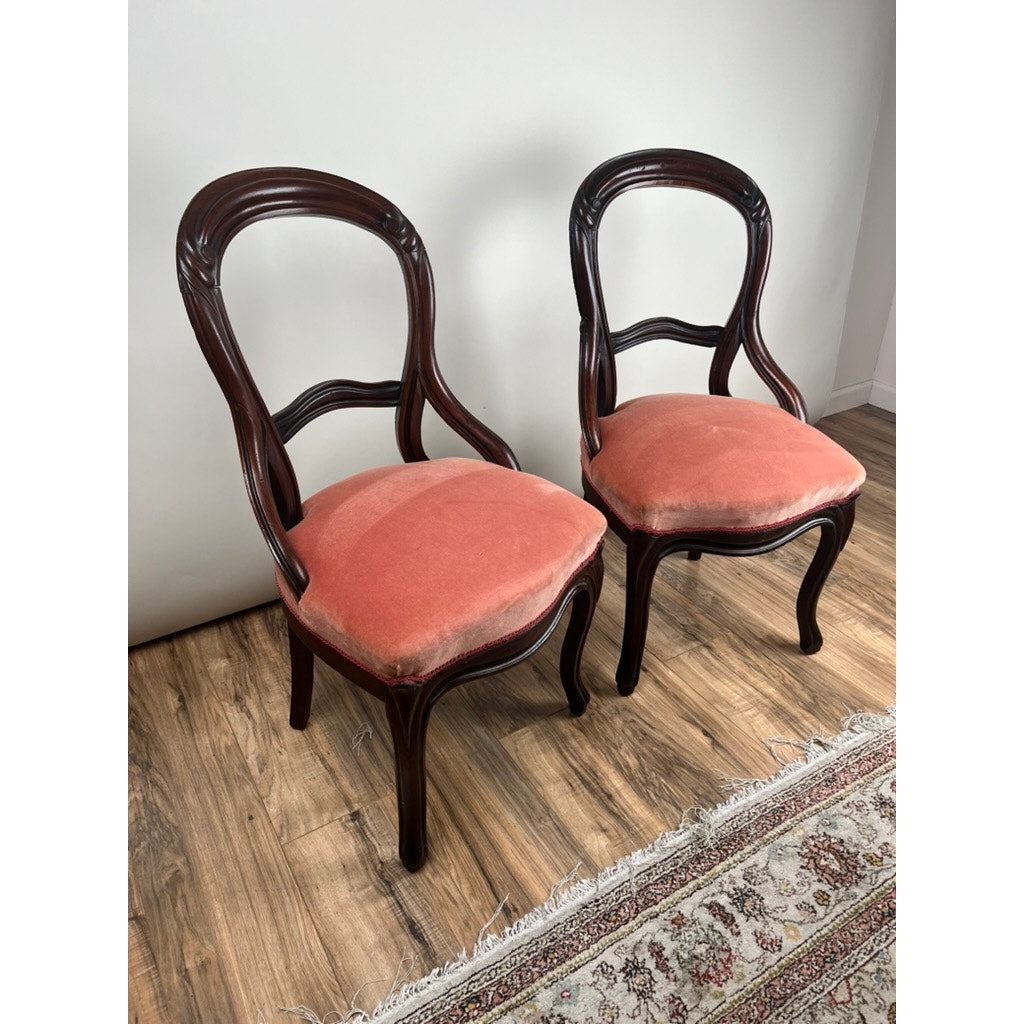 An Antique Pair of Victorian Walnut Balloon Back Side Chairs, featuring carved backs and vintage rose velvet upholstery, sit on a wooden floor beside a patterned rug and plain white wall.