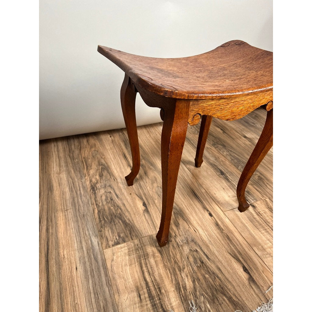 An Antique Arts and Crafts Saddle Shaped Footstool from c. 1900 sits on a wood-patterned floor against a plain white background, featuring curved legs and a slightly concave wooden seat. Brand: None/Unknown.