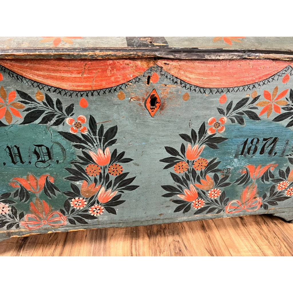 A close-up of an Antique Swedish Oak Kista Dowry Chest c. 1841, featuring floral designs in orange, green, and black on a distressed blue surface, shown sitting on a wood floor. Brand: None or Unknown.