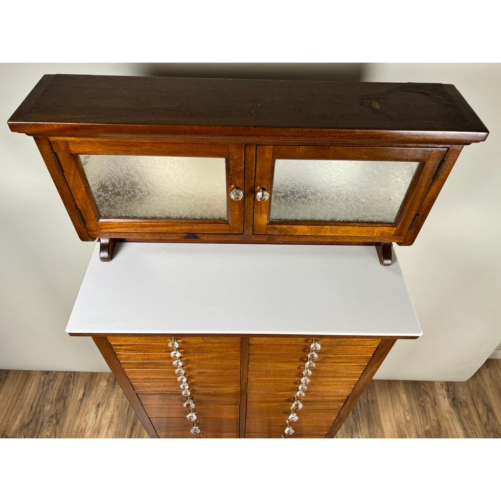 An Antique Mahogany Multi-drawer Dental Cabinet by The American Cabinet Co. (early 20th century) features frosted glass doors above a white countertop and crystal knob drawers, set on a wood-patterned floor.