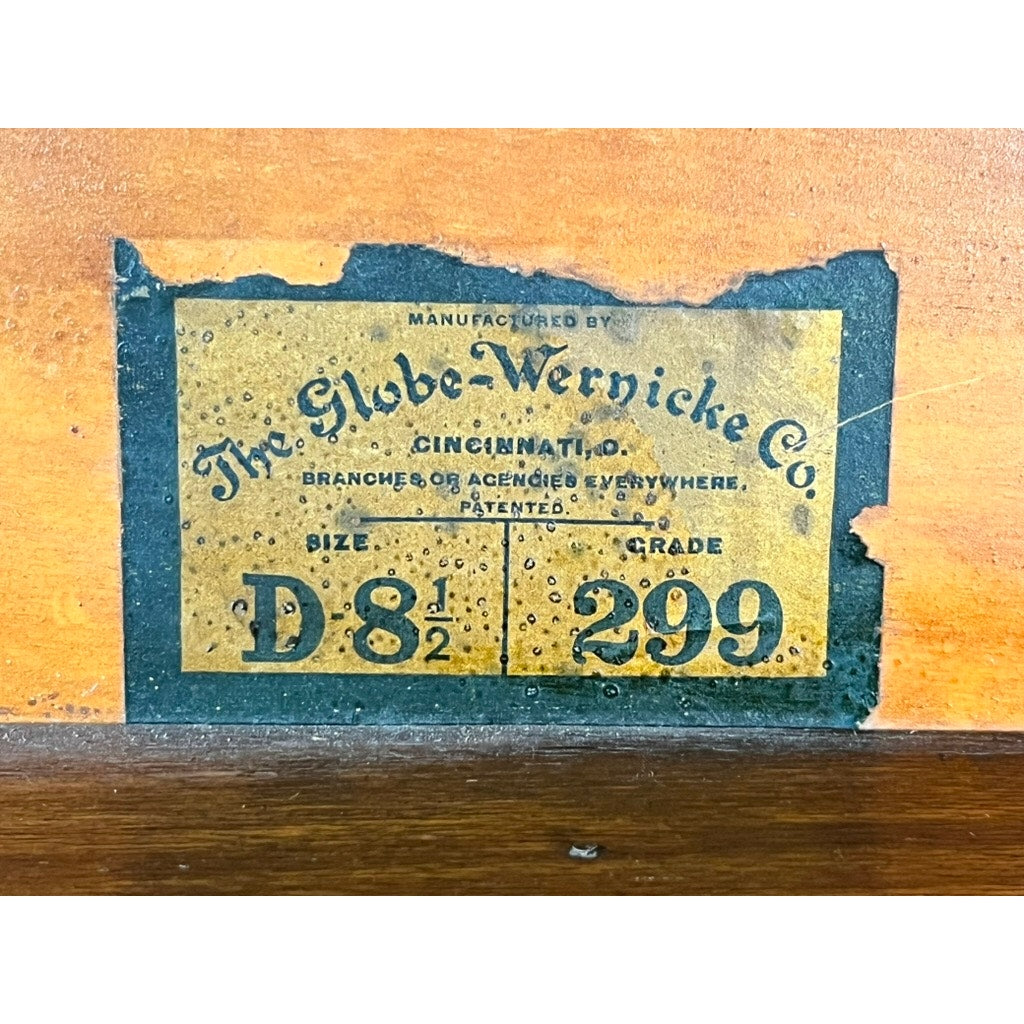 A weathered yellow label affixed to quarter-sawn oak reads: "The Globe-Wernicke Co. Cincinnati. Size D 8½, Grade 299." Displaying clear signs of age and wear along its edges, the label was originally applied to an Antique Globe-Wernicke Quarter Sawn Oak 6-Stack Barrister Bookcase Secretary Desk (c. 1905–1915) by Globe Wernicke, and now contrasts strikingly with the rich oak grain beneath it.
