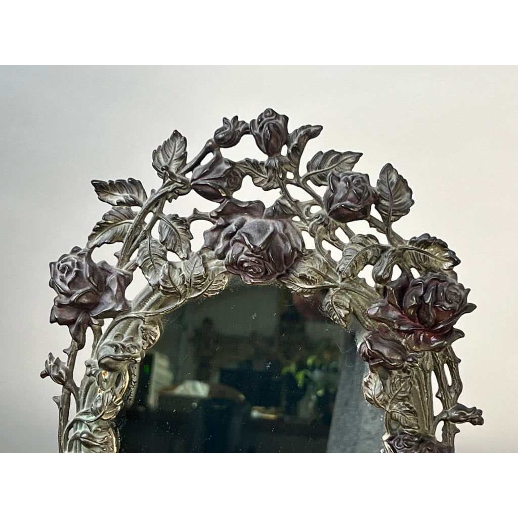 An Antique Art Nouveau Silvered Cast Brass Rose Vanity Dressing Mirror, dated 1909, features ornate rose and leaf designs on the upper edge, with a partial room reflection visible in the mirror glass.