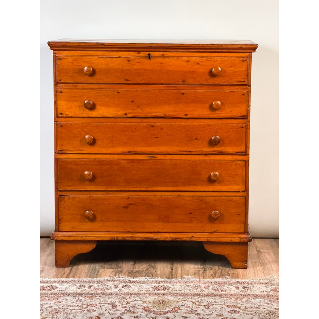 A Late 18th Century New England Pine Two-Drawer Blanket Chest by Pearl City Antiques, dating from c. 1780–1810, stands on a wooden floor beside a patterned rug, its timeless silhouette and patina evoking the charm of early American country furniture against a plain white wall.