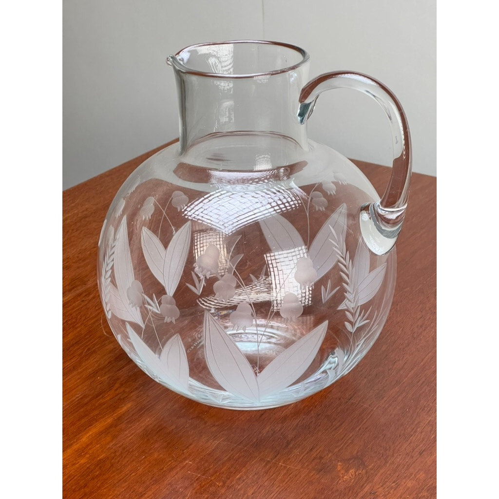A Vintage Tiffany & Co. Etched Crystal Pitcher, featuring a delicate lily of the valley floral motif, sits on a wooden surface against a plain light background.