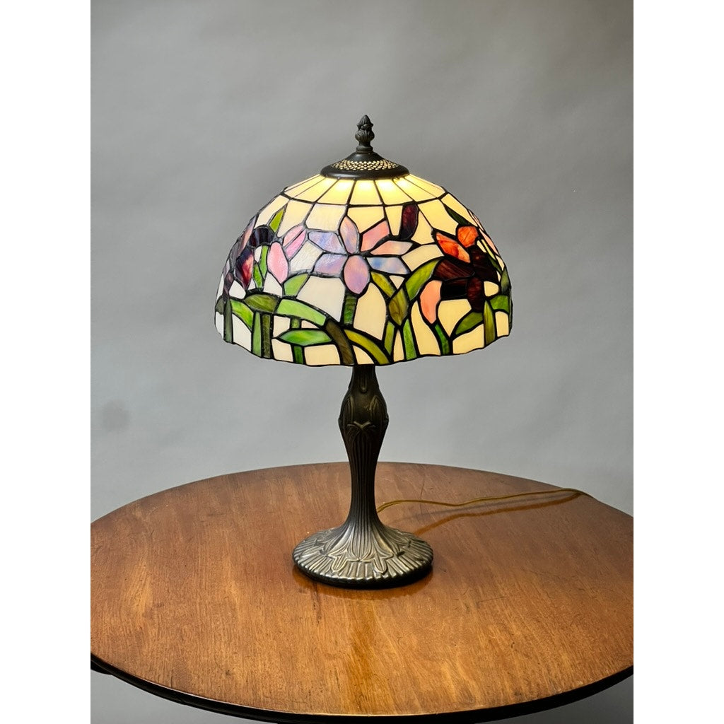 The Vintage Tiffany Style Floral Stained Glass Table Lamp, adorned with purple and red flowers and green leaves, rests on a round wooden table against a gray backdrop, showcasing classic elegance.