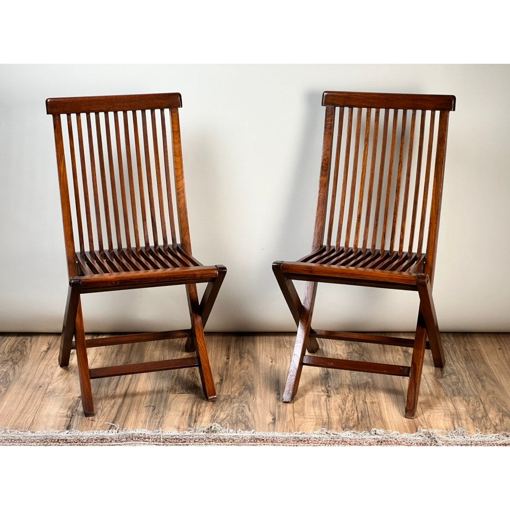 A Pair of Vintage Mid-Century Modern Style Teak Folding Chairs with vertical slats on the backrest and seat are placed on a wooden floor against a plain light-colored wall, with a rug partially visible at the bottom. Brand: Unknown.