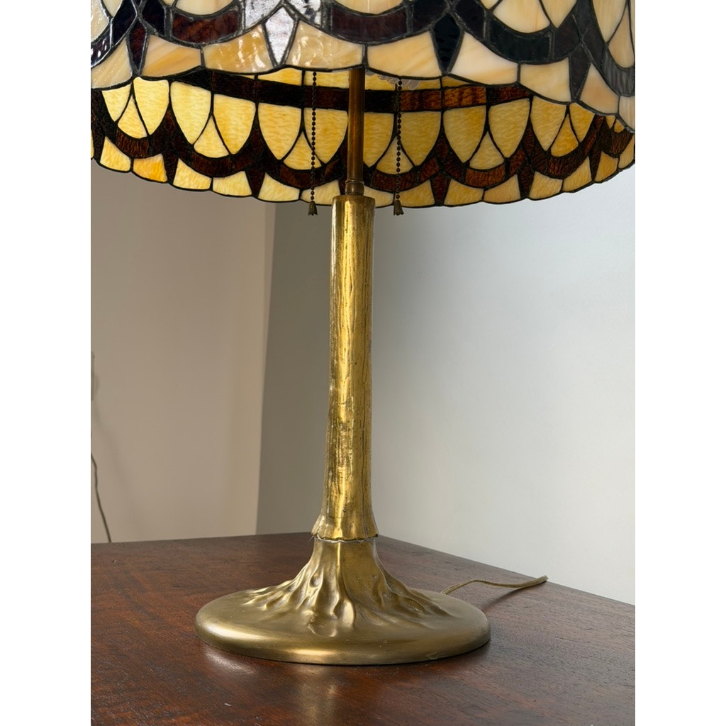 A Pearl City Antiques Vintage Tiffany Revival Leaded Stained Glass Table Lamp with a cast brass tree-form base sits on a wooden table against a light-colored wall, its cord visible on the surface.