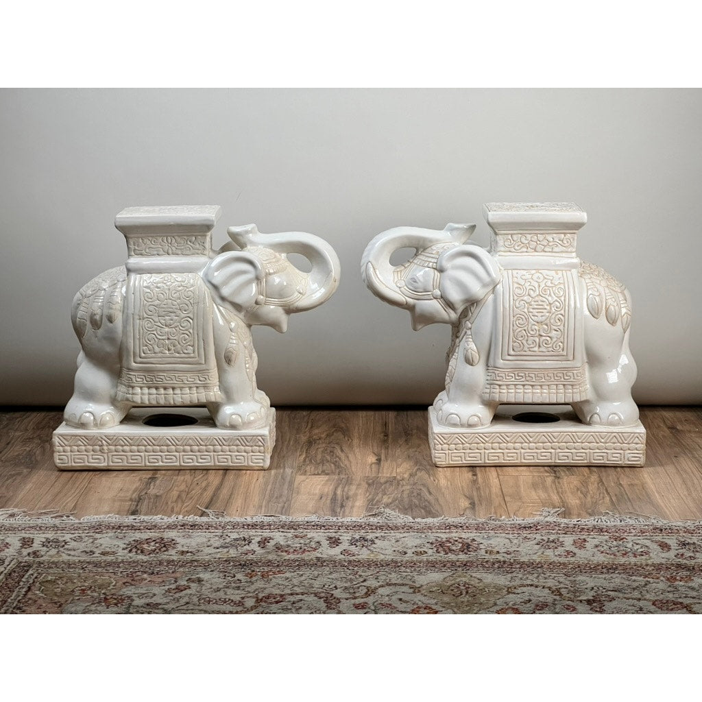 A Vintage Pair White on White Glazed Hollywood Regency Style Elephant Garden Seats stand side by side on a wooden floor, each featuring ornate patterns. A patterned rug appears in the foreground, enhancing the Hollywood Regency look.