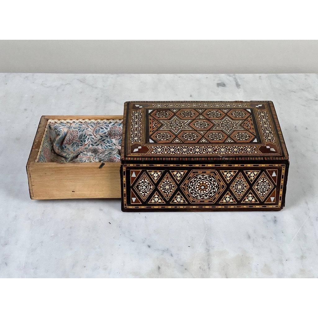 A late 19th-century Antique Persian (possibly Syrian) khatam inlaid wood secret drawer jewelry box sits open on marble, its ornate geometric patterns and faded fabric lining recalling the allure of a true antique.