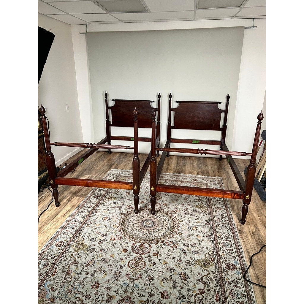 A Vintage Pair Centennial Solid Mahogany Twin Four Poster Beds c. 1940, without mattresses or bedding, are side by side on a patterned rug in a wood-floored room with a plain backdrop.