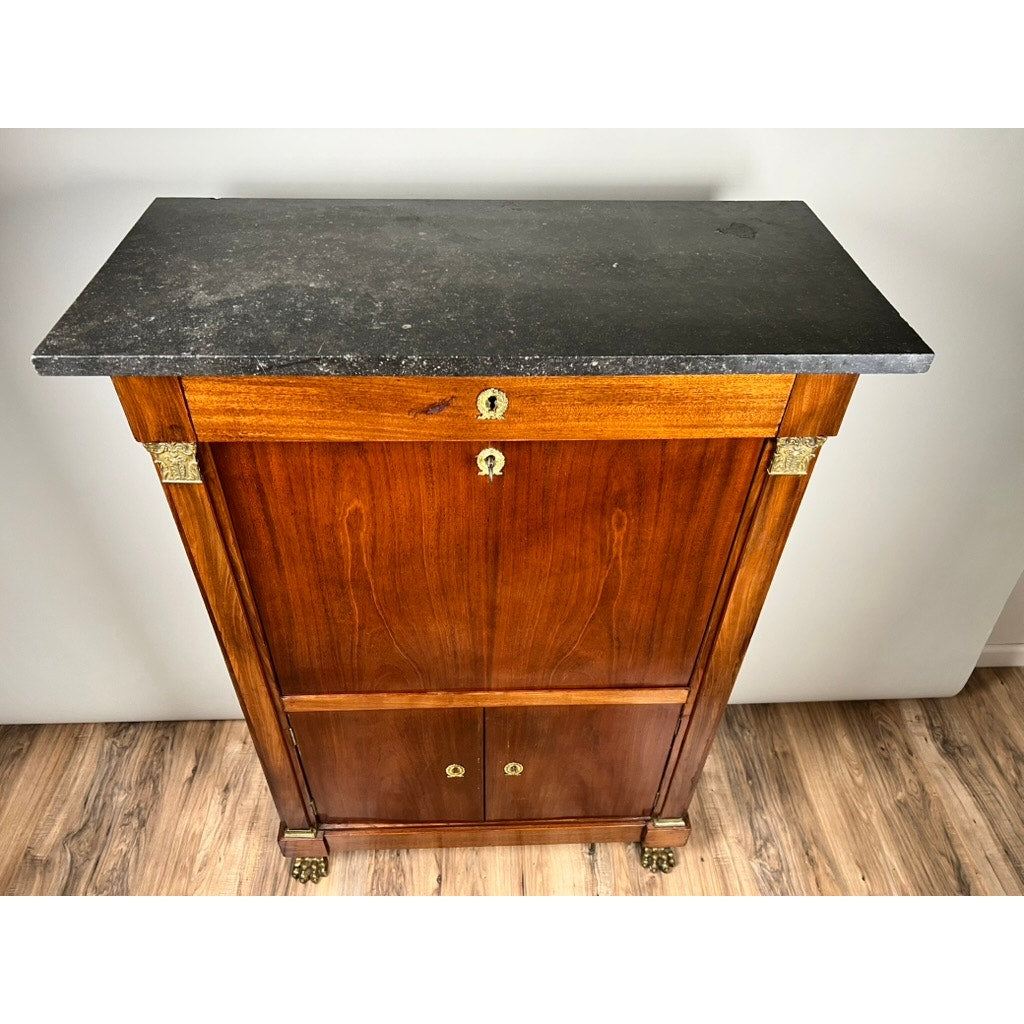 An Antique French Empire Secrétaire Abattant with mahogany veneer, ormolu mounts, lion’s paw feet, and a dark stone top stands on a wood floor. The mid-19th century piece features a keyhole drawer above two cabinet doors.