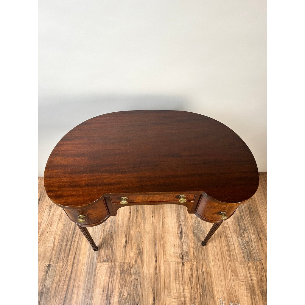 A Vintage Edwardian Sheraton Revival kidney-form dressing table (c. 1940) with mahogany veneers, curved front, two brass-handled drawers, and slender legs stands on a wooden floor against a plain light wall.