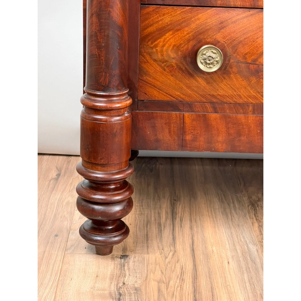 Close-up of a turned wooden leg on an Antique American Empire Albany Four Drawer Chest c. 1840, featuring a round brass knob and flame mahogany finish, shown on a wooden floor. Brand: None/Unknown.