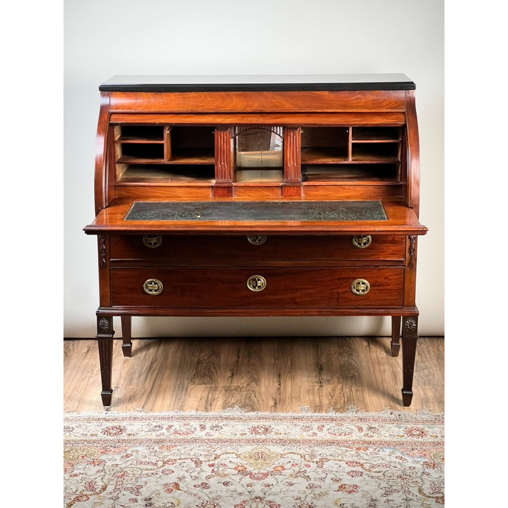 An Antique Directoire Style French Cylinder Desk with a slate top, c. late 19th century, featuring a roll-top, glass-front cabinet, small shelves, and three brass-handled drawers is displayed on a wooden floor with a decorative rug.