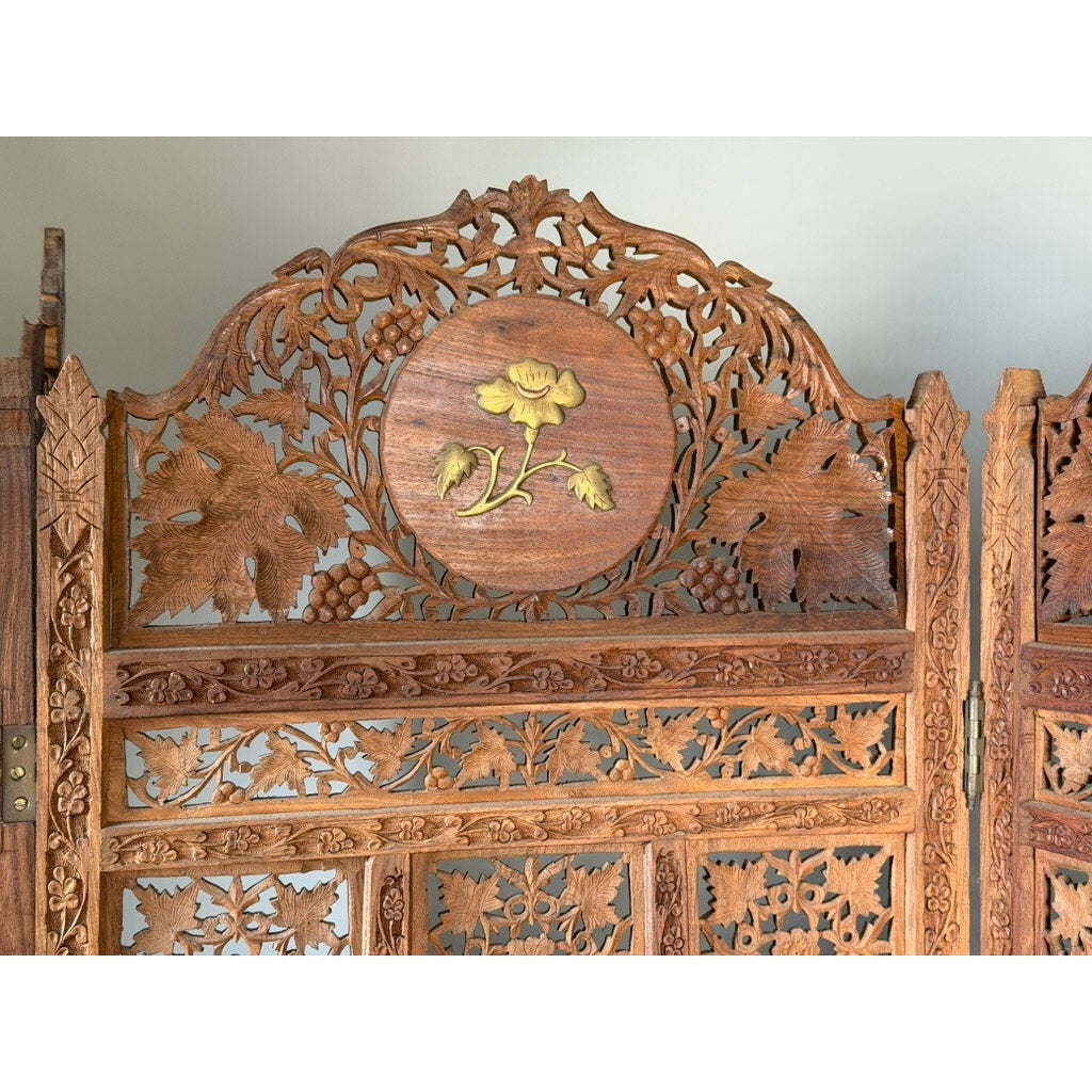 Early 20th Century Anglo-Indian Carved Teak Four-Panel Screen from Pearl City Antiques, featuring intricate floral and leaf carvings with applied brass floral decoration. The top showcases a circular centerpiece adorned with a raised yellow flower design, surrounded by detailed latticework of leaves and vines, evoking the elegance of an antique decorative screen.