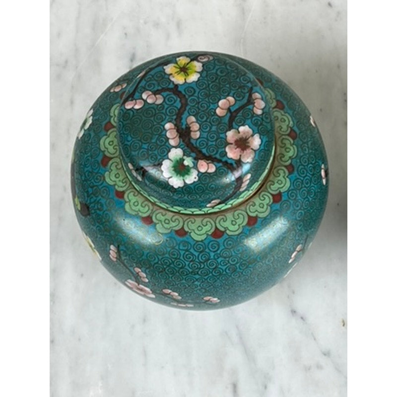 A vintage pair of Chinese cloisonné ginger jars with lids, featuring enamel designs of pink cherry blossoms and green swirling clouds, displayed on a white marble surface. Brand unknown.