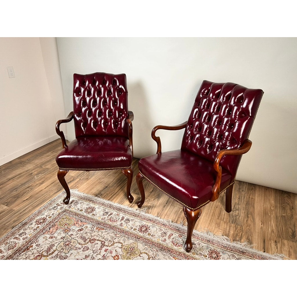 A Pair of Chippendale Style Tufted Burgundy Leather Library Armchairs with elegant wooden frames sit atop a patterned rug in a room with light-colored walls and wood flooring.