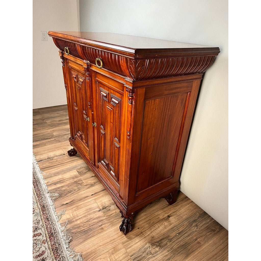 An Antique English Jacobean Revival Mahogany Buffet (c. 1915–1925) with ornate carving, brass handles, and ball-and-claw feet stands against a light wall on wood flooring beside a patterned rug.
