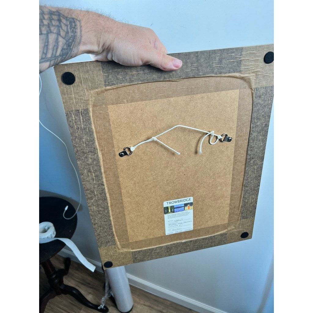 A hand holds the back of a Trowbridge Vintage Custom Frame for the “Phlox” botanical print after Louis van Houtte, showing four black corner pads, a hanging wire, a bottom center label, and part of a table with rolled paper in the background.