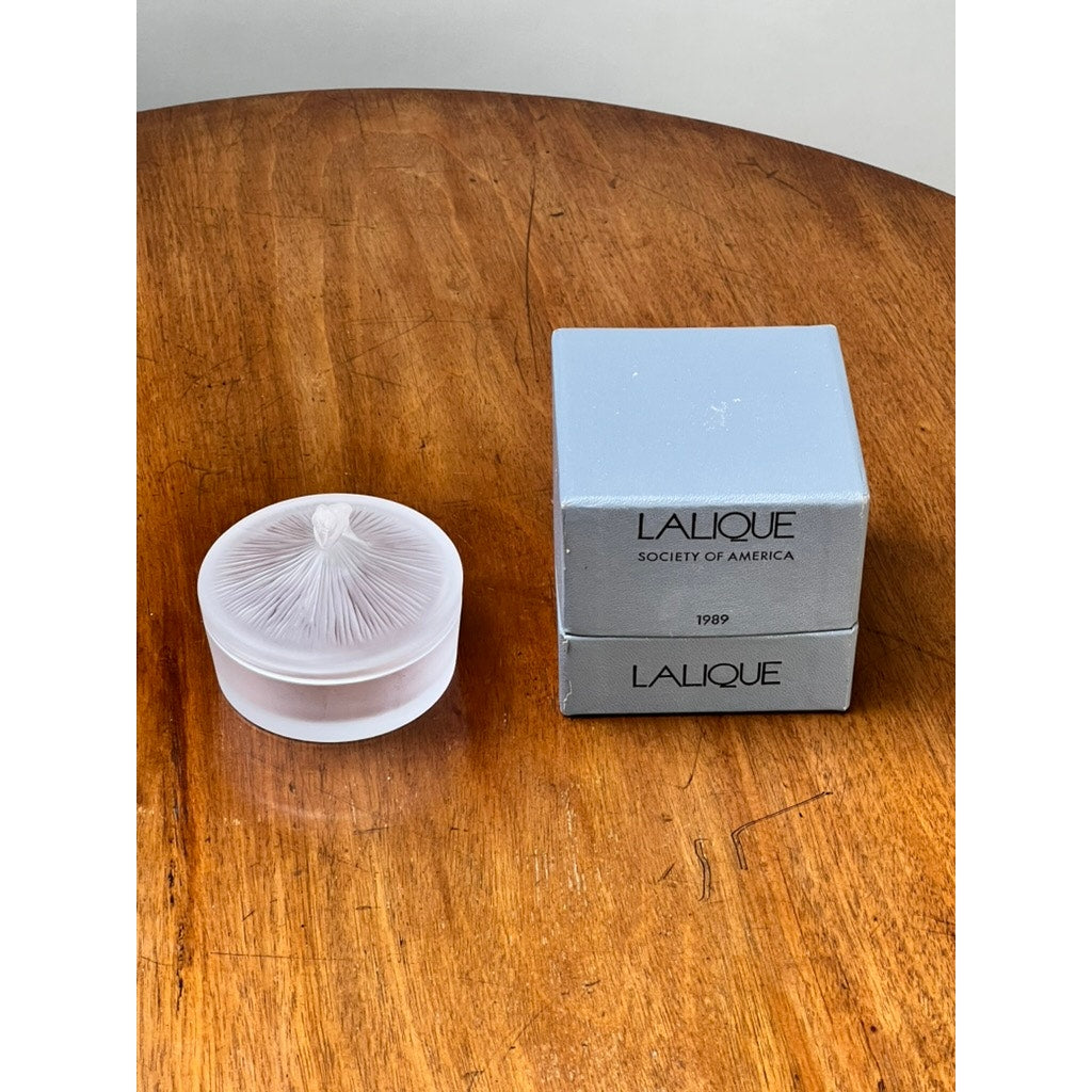 A Vintage Lalique France Ballerina Frosted Crystal Covered Powder Box sits next to two gray Lalique Society of America 1999 boxes on a wooden table.