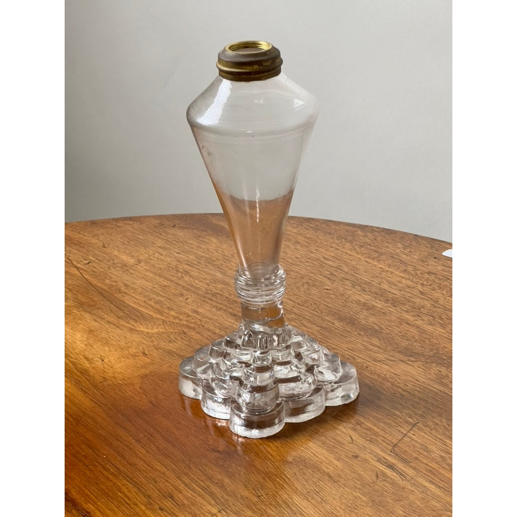 An Attributed Boston & Sandwich Pressed Glass Boudoir Fluid Lamp, circa 1850, featuring a vintage clear glass base with a brass collar and a tiered geometric foot reminiscent of mid-19th century design, sits on a wooden table against a plain light background.