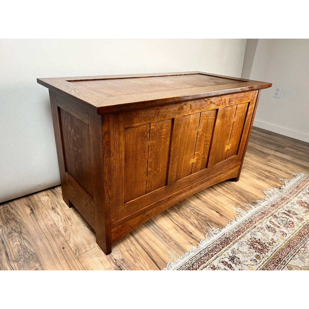 A Stickley Vintage Harvey Ellis Millennium Edition Oak Blanket Chest with paneled detailing, finished in rich brown, sits on a wooden floor by a patterned rug and a white wall.