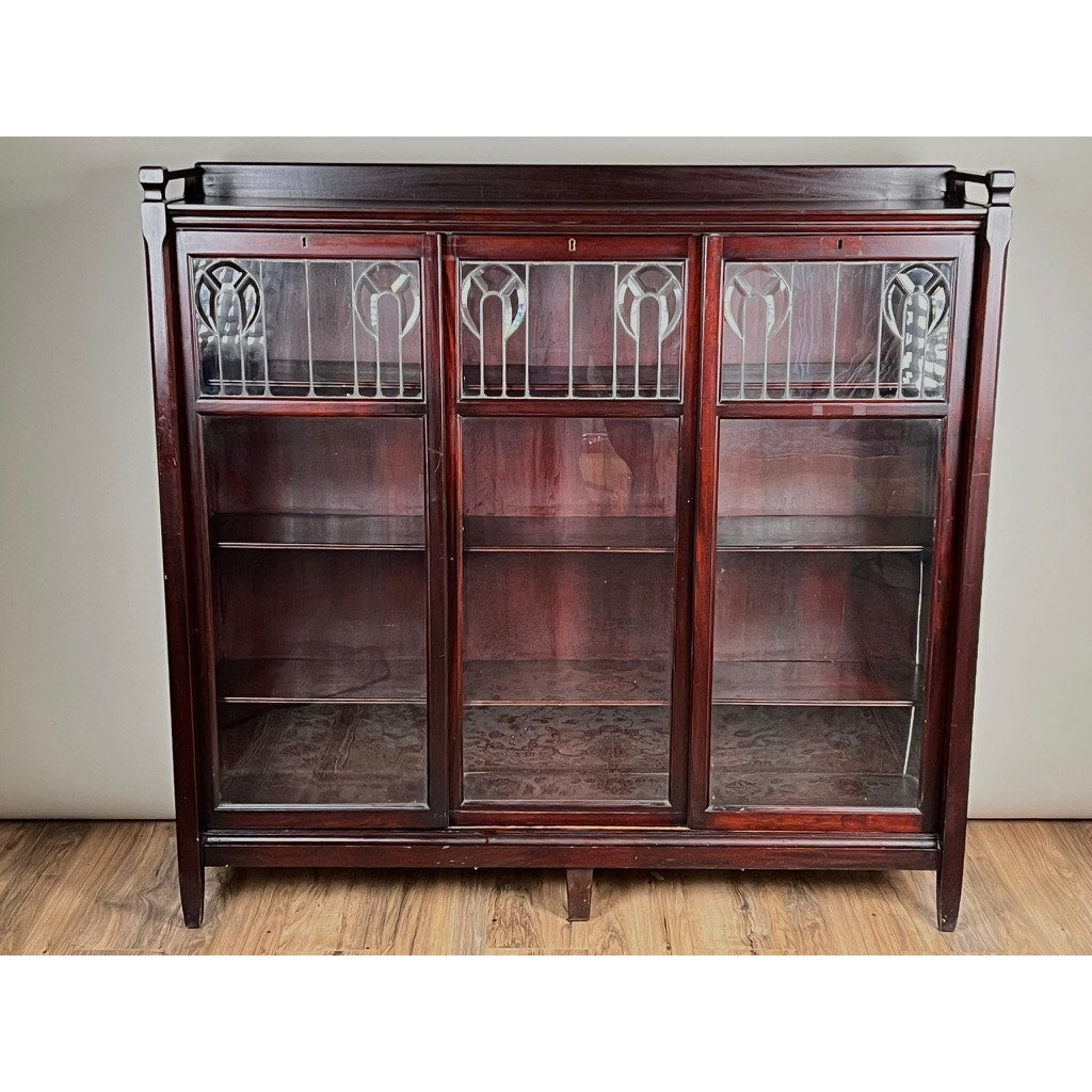 An Antique Arts & Crafts Leaded Glass Mahogany Bookcase c. 1920 with three leaded glass doors, decorative panels at the top, and three shelves stands on a wooden floor against a plain light-colored wall.