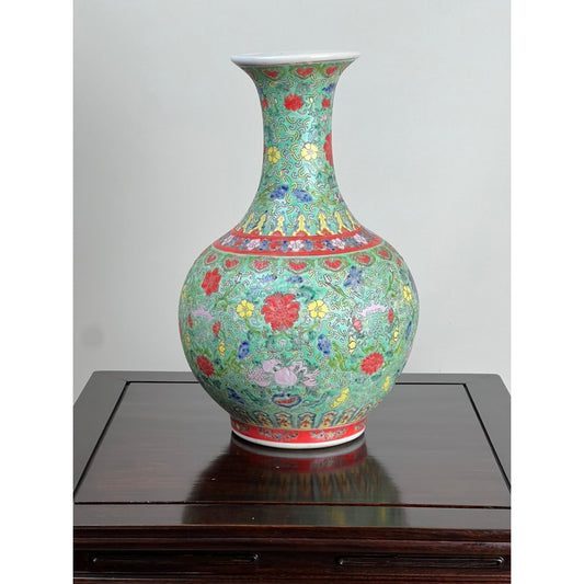 An Antique Qing Dynasty Famille Rose Chinese Porcelain Vase with Qianlong Marking from Pearl City Antiques, featuring a long neck and colorful red, yellow, and blue floral patterns on a green background, is elegantly displayed on a dark wooden table.