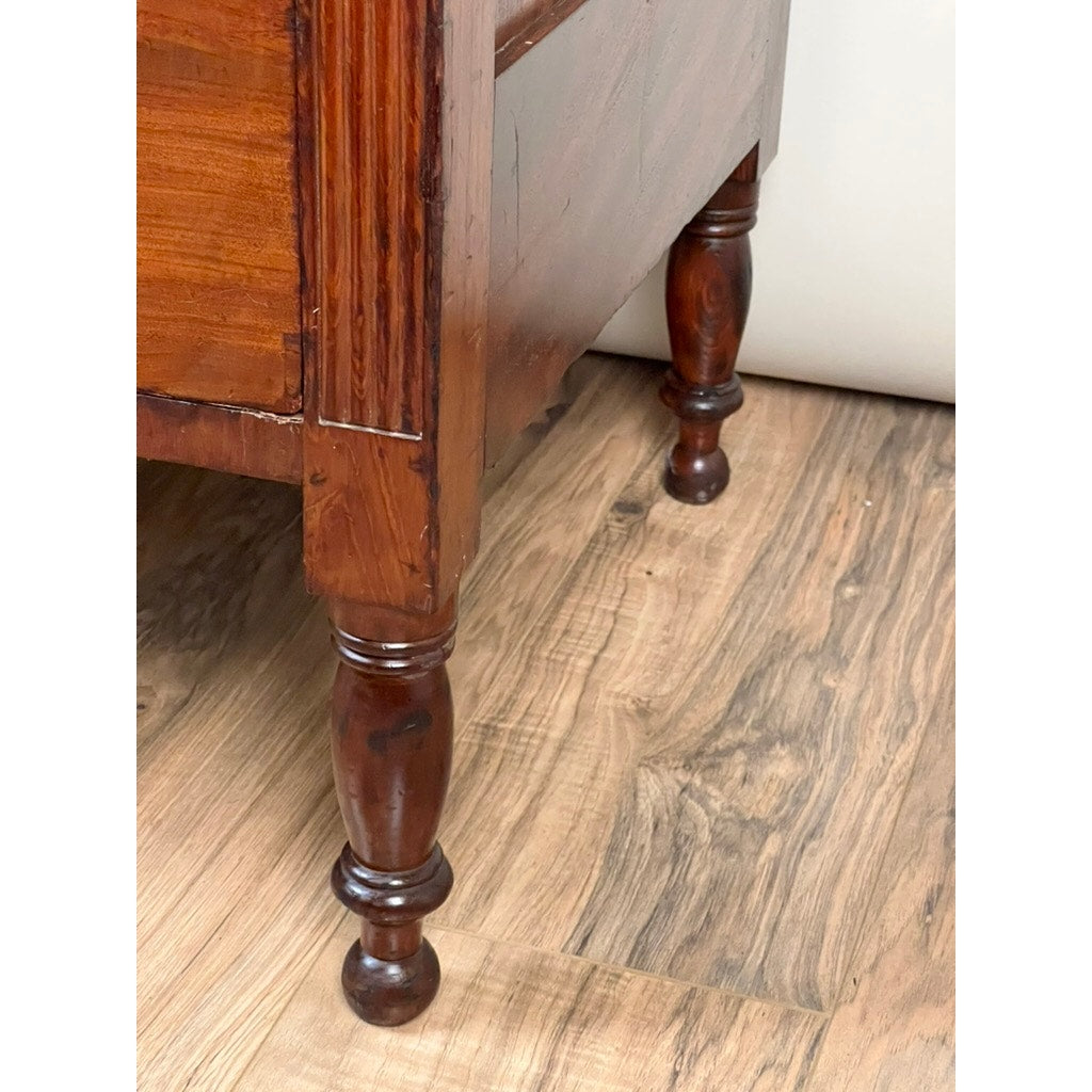 Close-up of the corner of an Antique George III Sheraton Flame Mahogany Chest of Drawers (Early 19th Century) with ornate, turned legs, shown on a wood-patterned floor. Brand: Unknown.