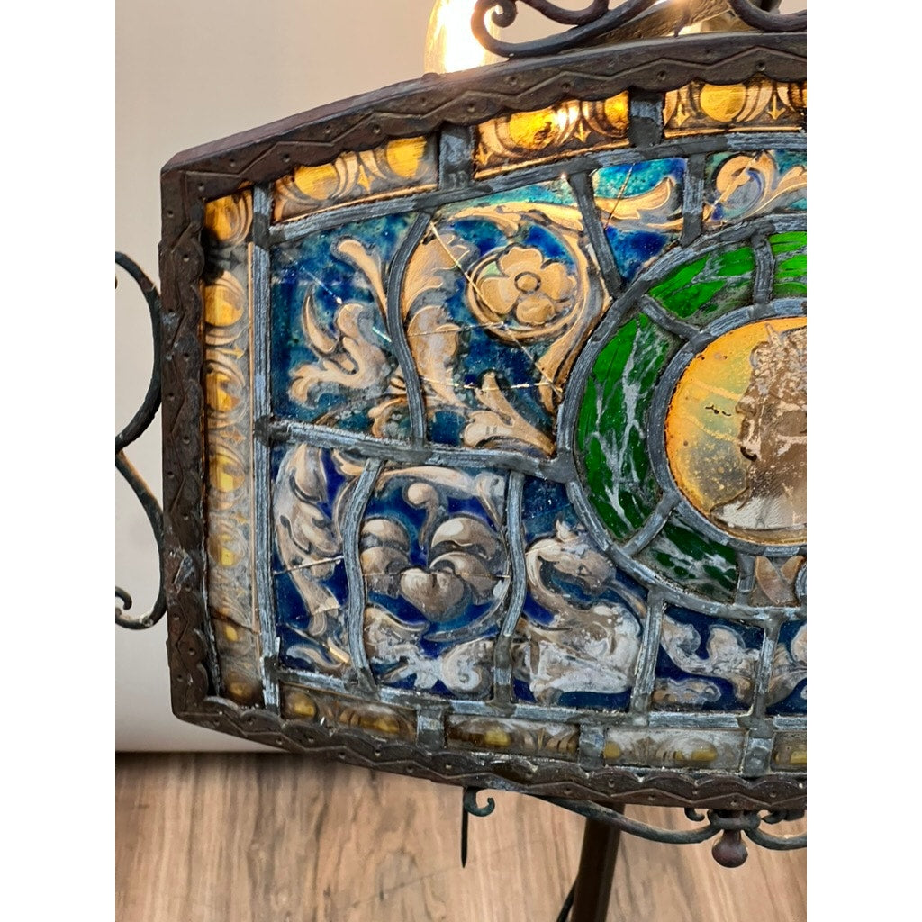 A close-up of an Antique Five Burner Lucerne Whale Oil Lamp with Napoleon I Fireplace Screen Shade, showcasing ornate blue, yellow, and green floral stained glass in a decorative metal frame against a wooden floor and neutral wall.