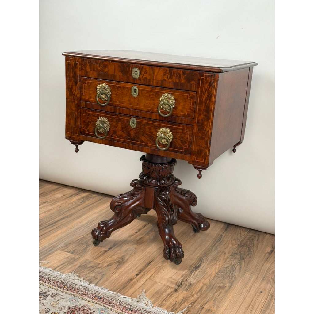This American Classical Figured Mahogany Work Table, circa 1830–1845 features two drawers with brass lion-head pulls, supported by a carved pedestal base with four claw feet on casters.
