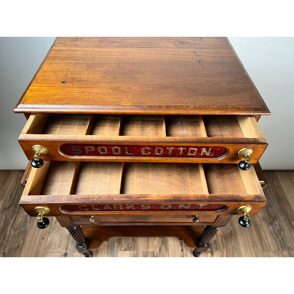 An Antique Victorian Pine Two Drawer Clark’s O.N.T Spool Cabinet by Clark's ONT, c. late 19th century, with labeled drawers, multiple compartments, and brass knobs stands on a wood floor.