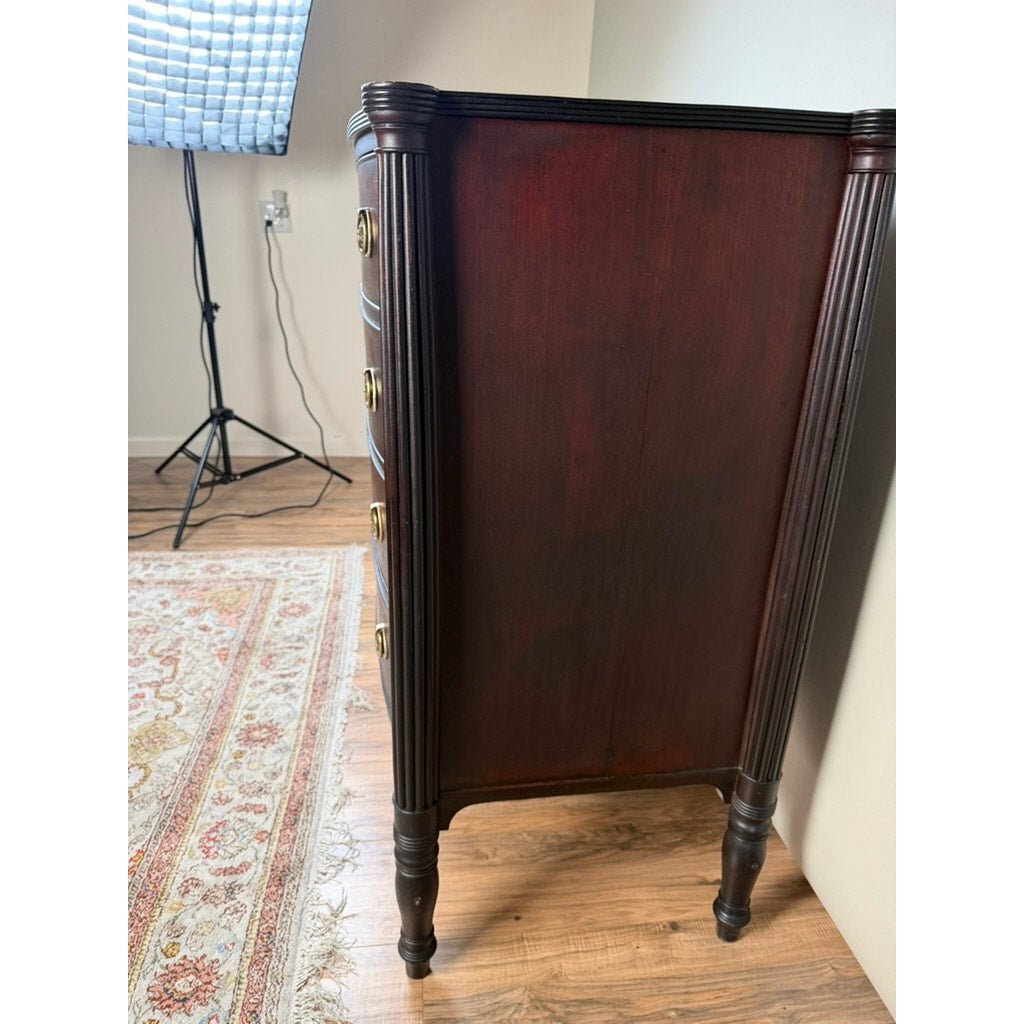 An Early 19th Century New England Federal Mahogany Bow-Front Chest of Drawers (c. 1810–1825) is positioned against a wall; its elegant brass handles highlight the classic Federal style. In the background, a patterned rug and a photography light with a softbox can be seen on the wooden floor.