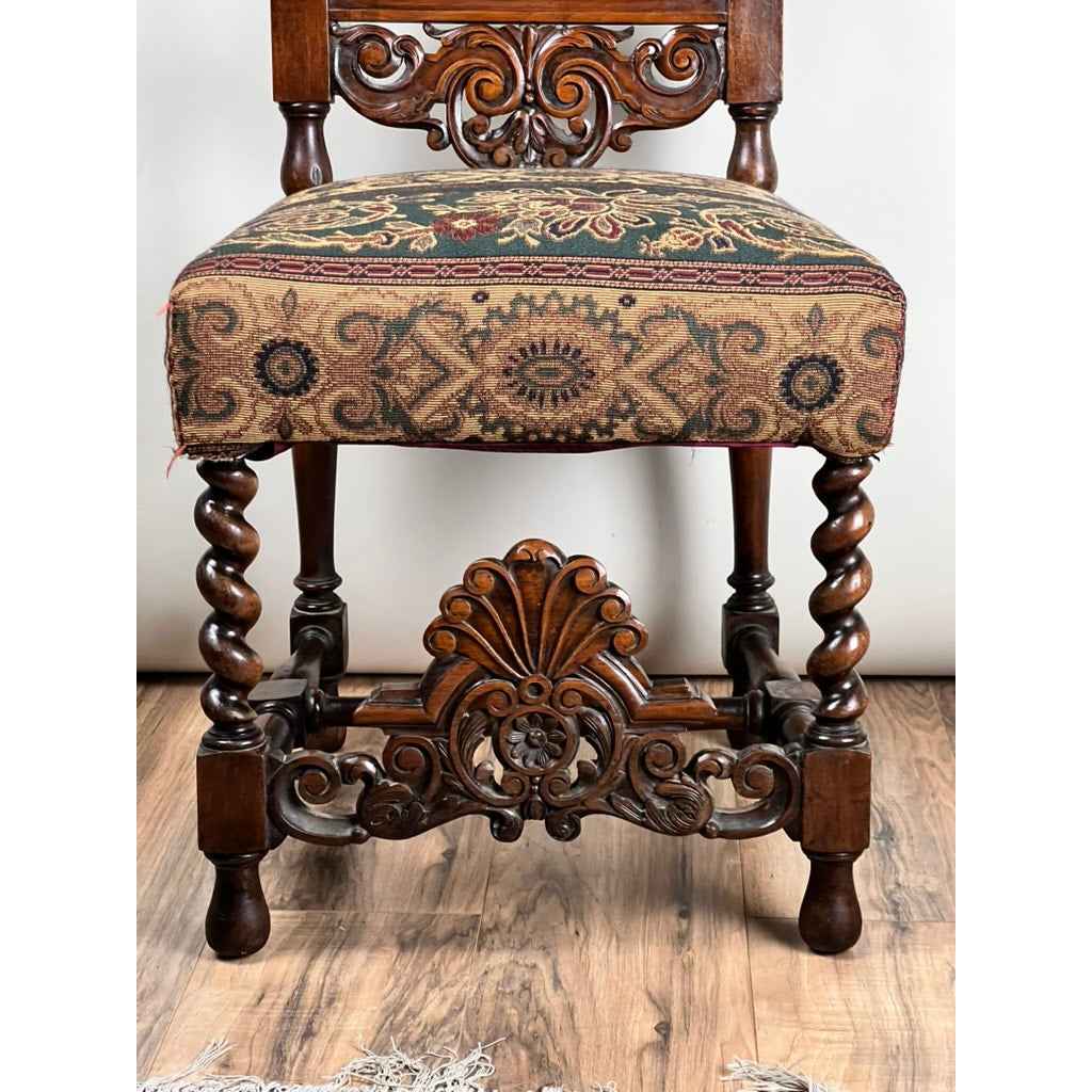 An Antique Carolean Revival Side Chair from the late 19th century, featuring intricate carvings, twisted legs, and a green, red, and gold patterned upholstered seat, is displayed on a wooden floor. Brand: Unknown.