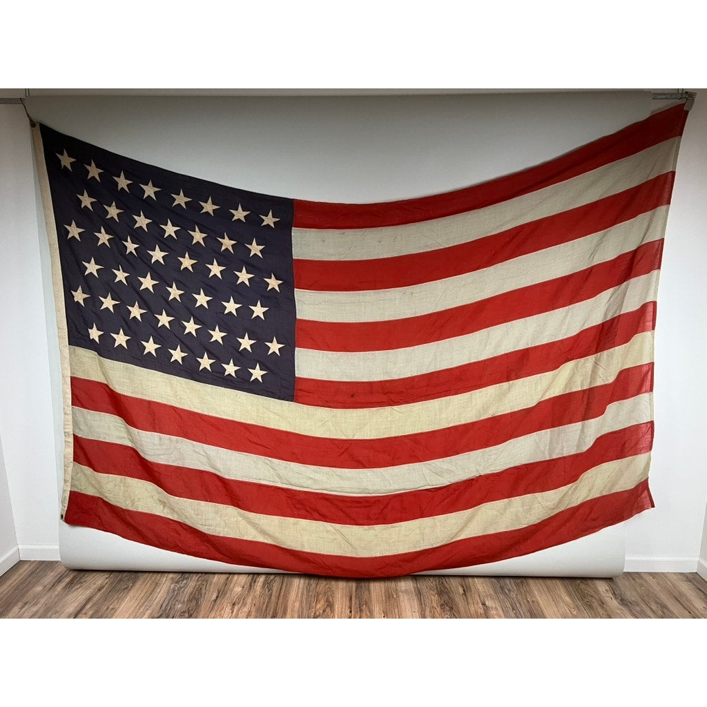 An antique 45 star American flag from the Spanish–American War era (9’9" x 6’) is displayed hanging on a white wall above a wooden floor, adding vintage Americana charm. Brand: Unknown.