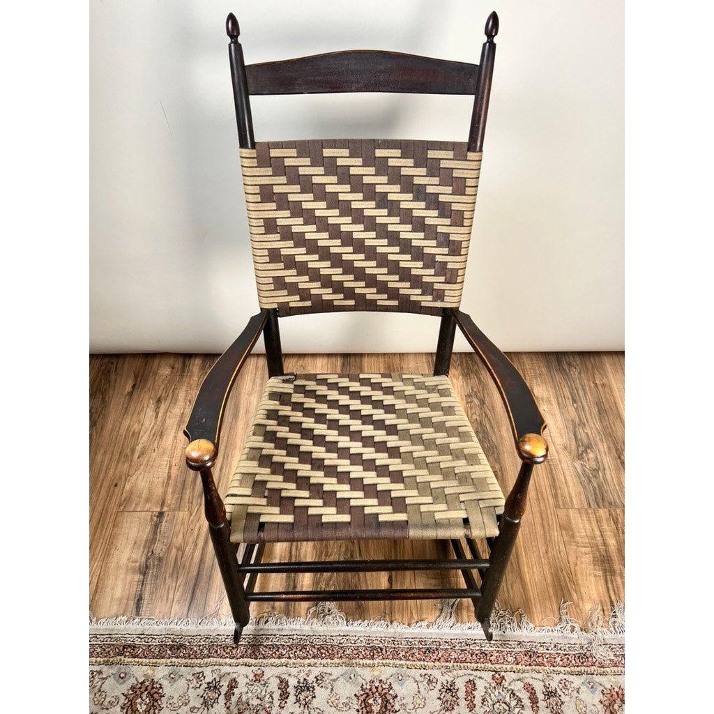 An Antique Shaker #6 Tape Seat Rocker by Mt. Lebanon Shakers, late 19th century, with a brown and beige geometric woven tape seat and back, sits on a wooden floor next to a patterned rug.