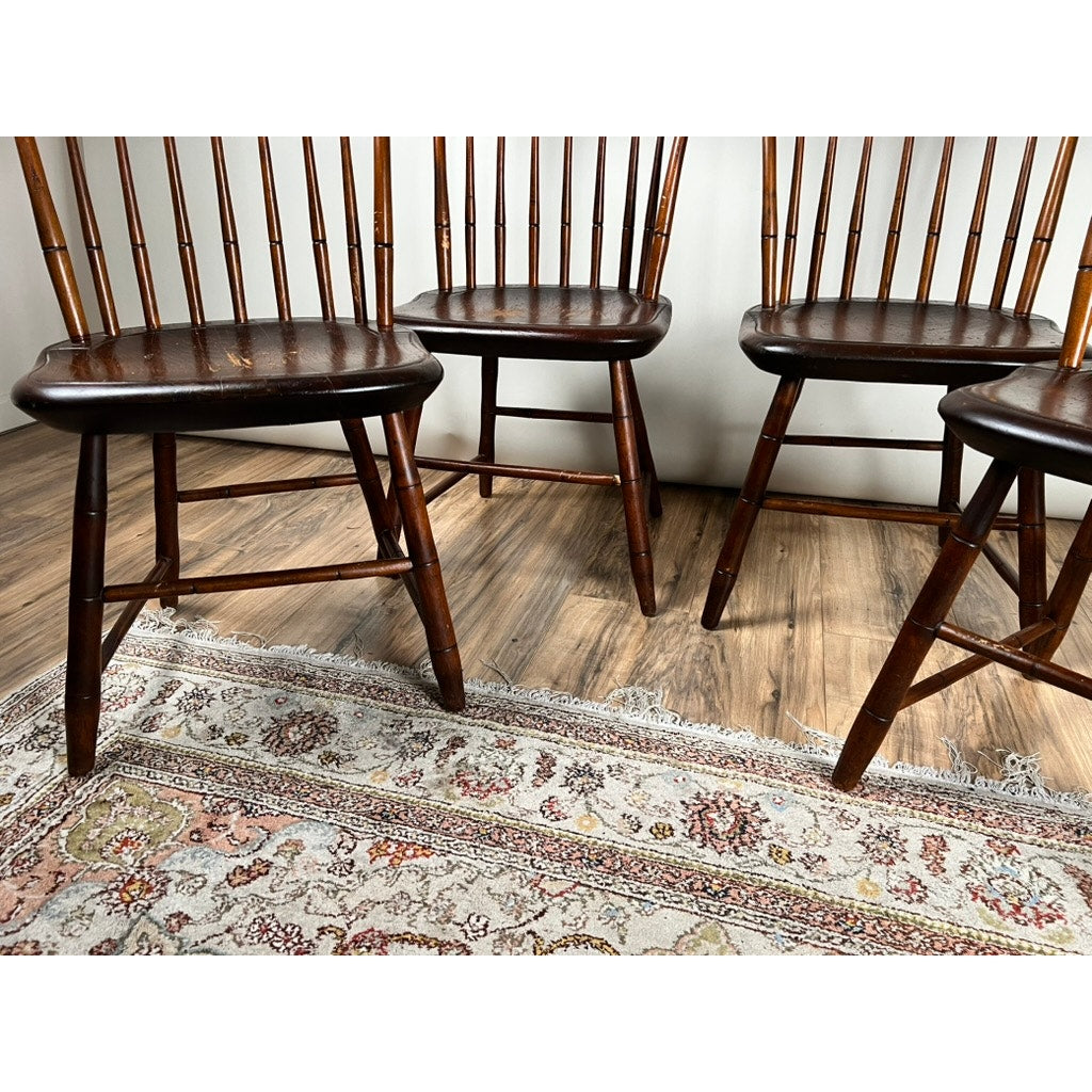 A set of four early 19th-century New Hampshire Step Down Windsor Chairs by JR Hunt, each with dark seats, are placed on a patterned rug atop wood flooring against a white wall, showing only their lower halves.