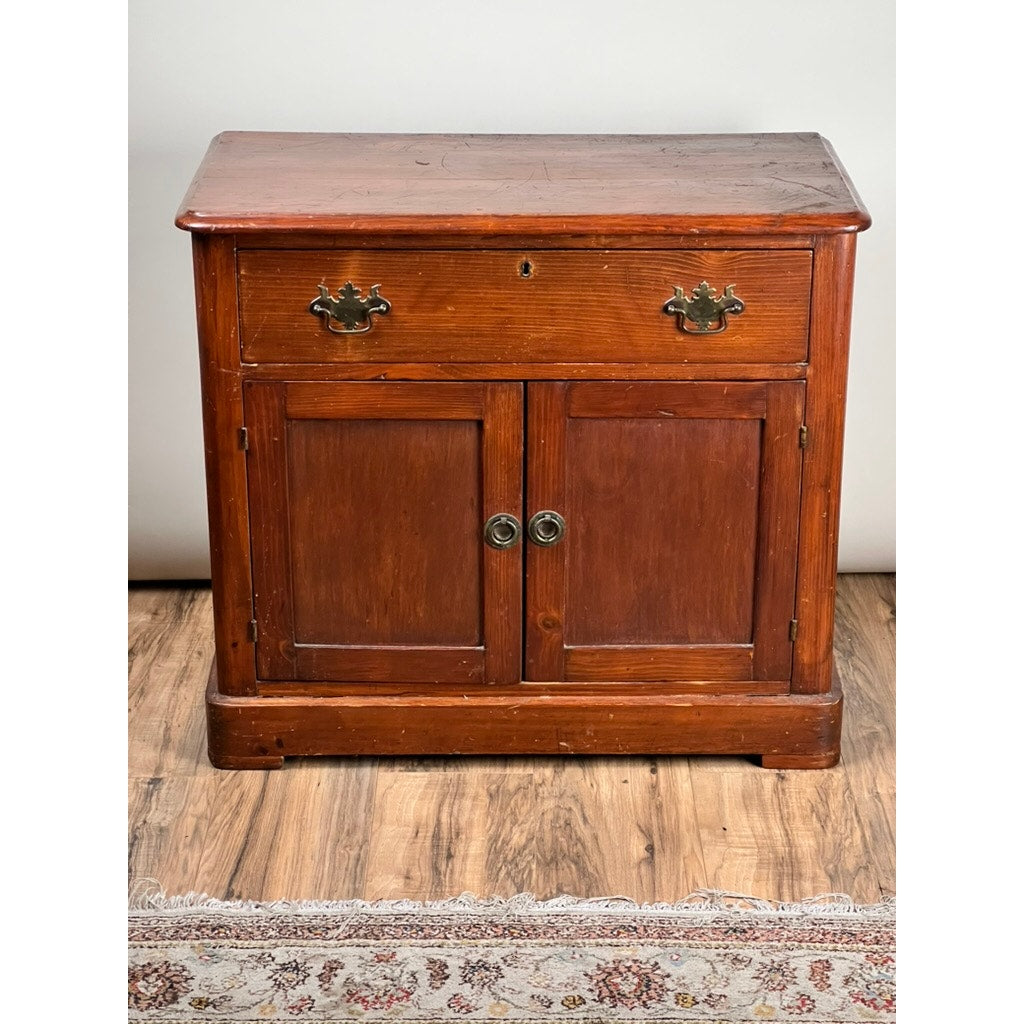 An Antique Victorian Pine Commode c. 1900 with a top drawer, two doors below featuring round metal handles and brass hardware stands on a wooden floor beside a patterned rug—ideal vintage bedroom storage furniture.
