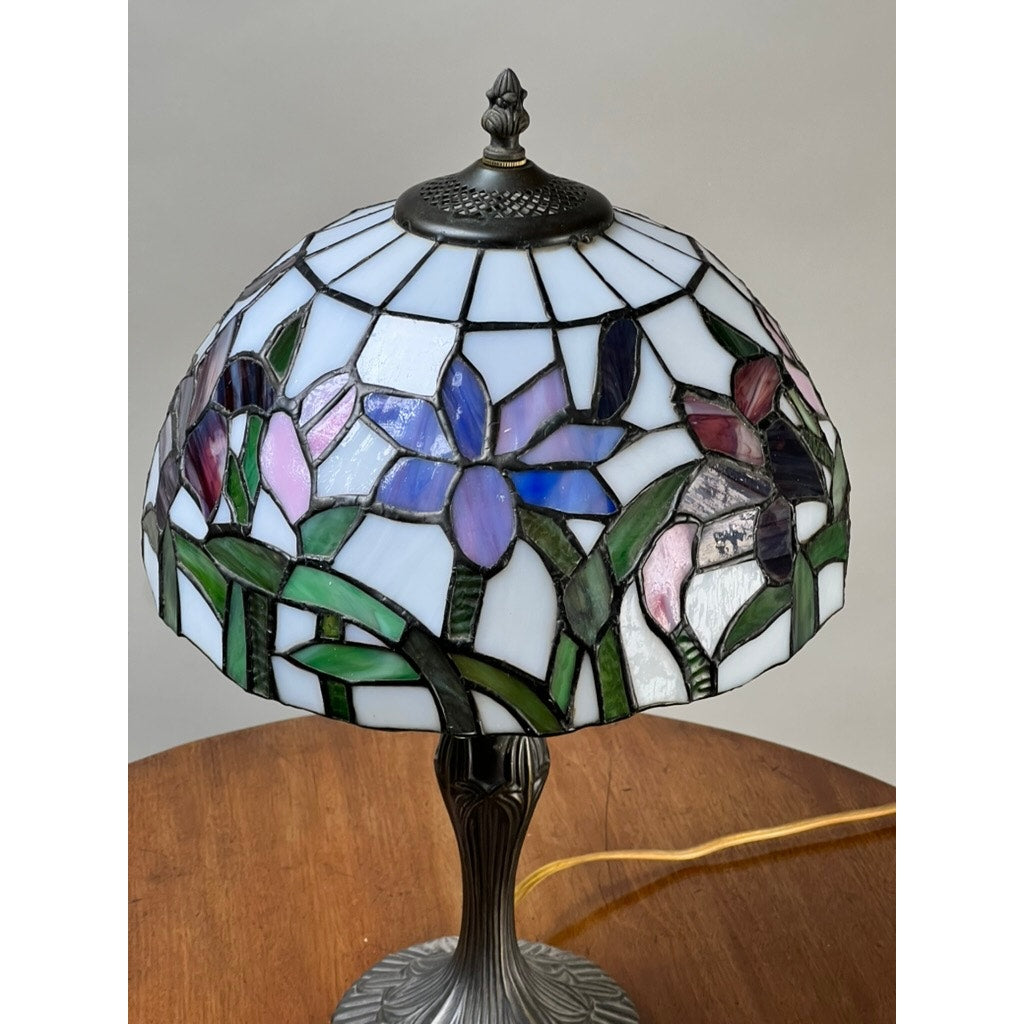 A Vintage Tiffany Style Floral Stained Glass Table Lamp with blue, purple, pink, and green floral patterns sits on a wooden surface. The lamp features a decorative metal base and is turned off. Brand is none or unknown.