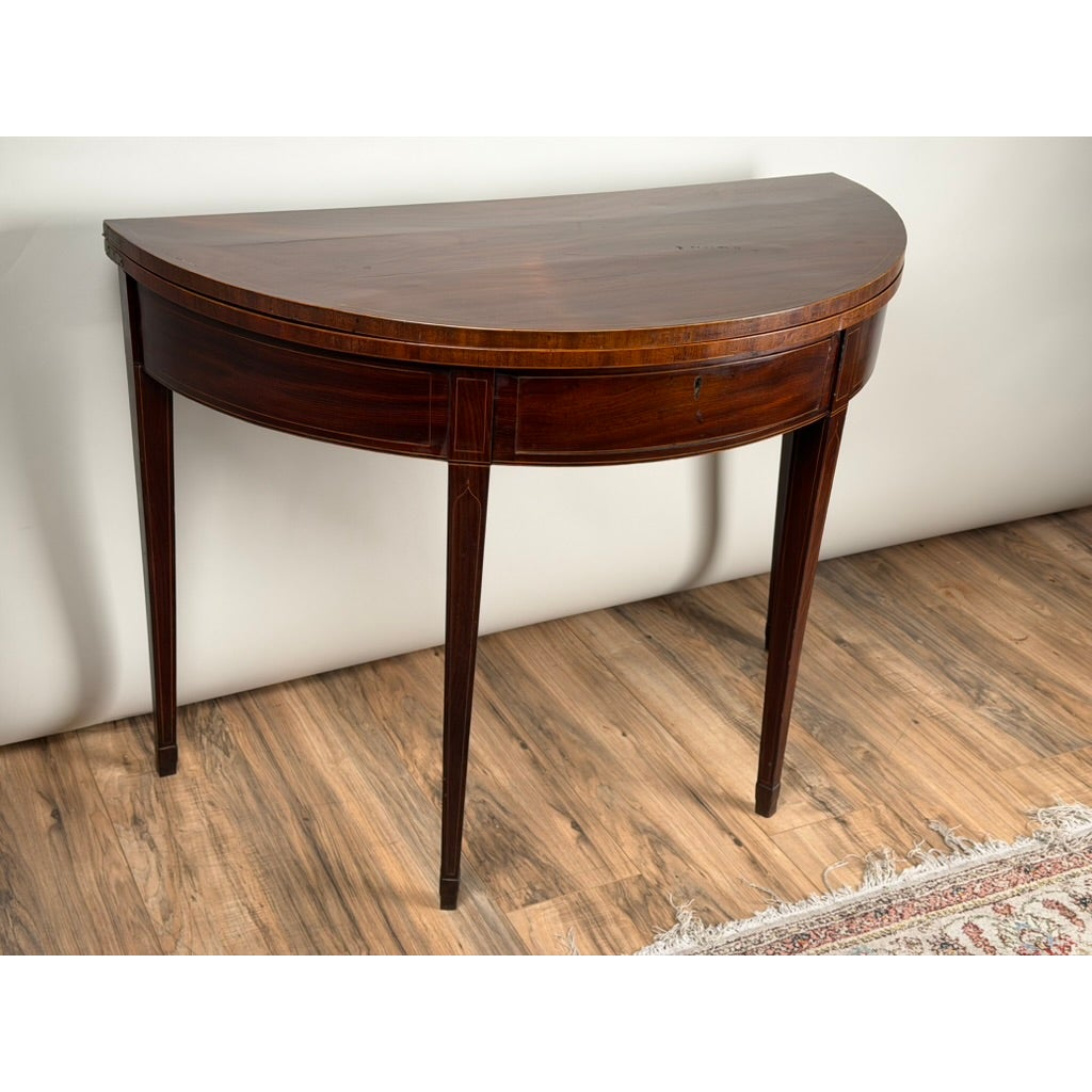 An English George III Mahogany and Satinwood-Inlaid Demilune Games Table, circa 1790–1810, featuring a mahogany demilune shape with four tapered legs and inlaid detailing along the front apron.