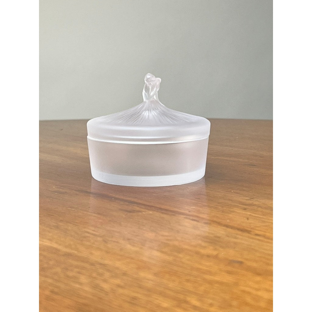 The Vintage Lalique France Ballerina Frosted Crystal Covered Powder Box by Lalique, featuring a rounded ribbed lid with a decorative knob, sits on a wooden surface against a plain light background.