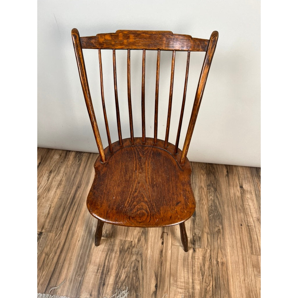 An Antique Step Down Windsor Chair in oak and pine (early 19th century) with a curved seat, vertical slat backrest, and angled legs sits on a wood-patterned floor against a plain light-colored wall.
