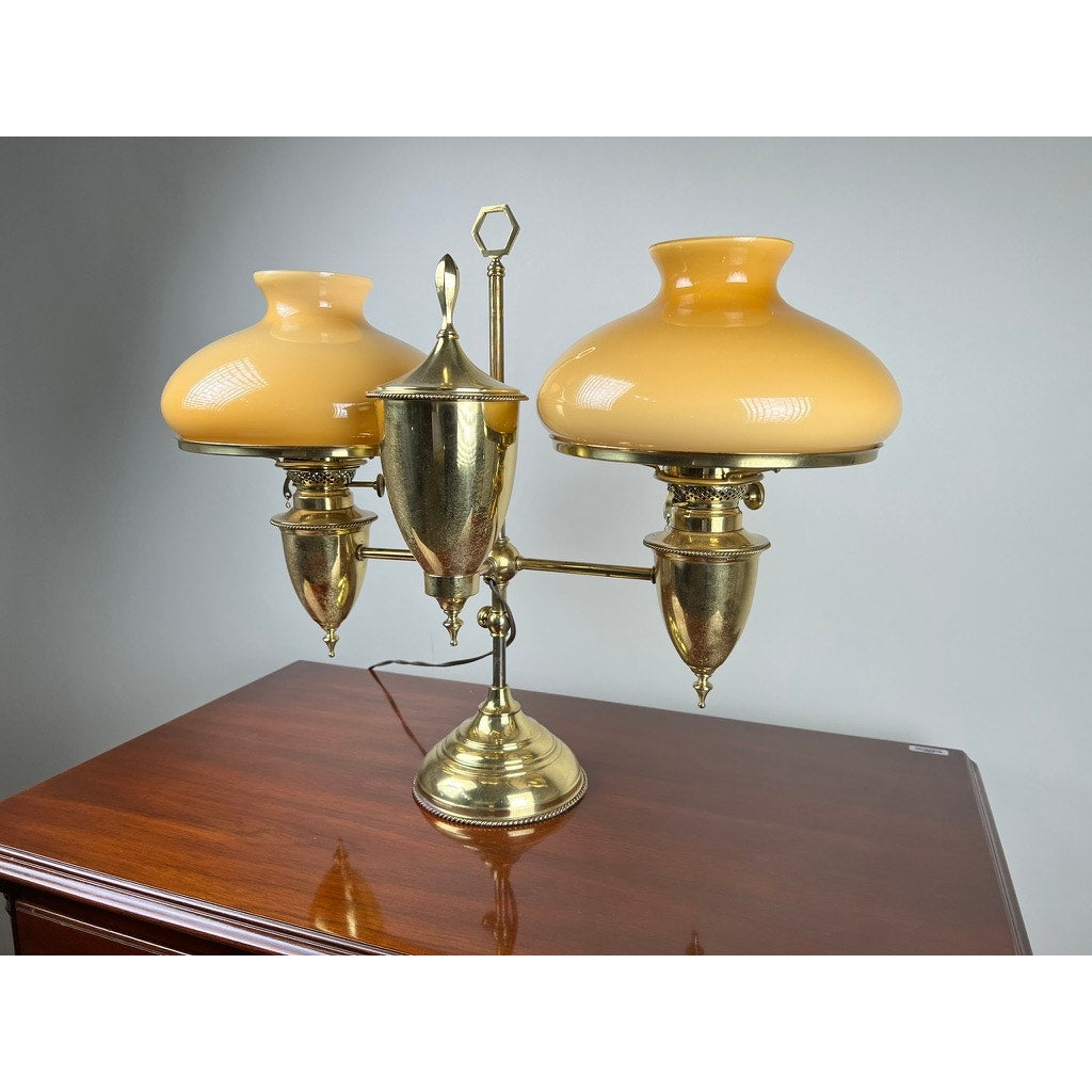The Antique Bradley and Hubbard Brass Double-arm Student Lamp with caramel shades features ornate detailing, a central finial, and sits on a polished wooden surface against a plain gray background.