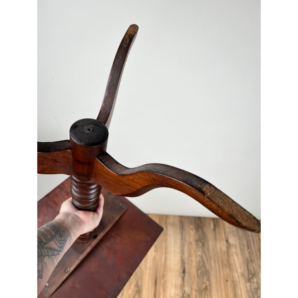 A tattooed arm holds the base of an Antique Low Cherry Candle Stand c. Mid 19th Century with three curved legs, set on a wooden floor against a plain white wall. Brand: Unknown.