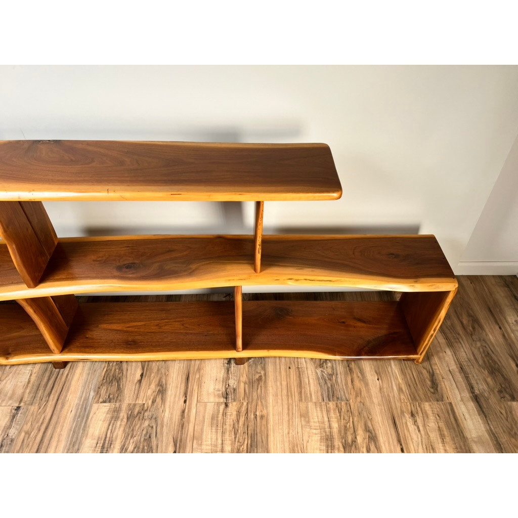 The Vintage Mid-Century Modern Style Live Edge Walnut Bookshelf features two tiers and an asymmetrical natural edge design, adding style against a white wall and wood-patterned floor. Brand name: none/unknown.