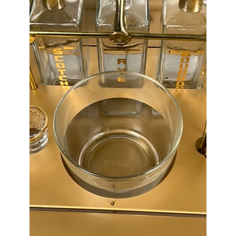 A clear glass bowl sits on a gold surface next to a Vintage Mid Century Modern Bar Caddy c. 1960s holding three decanters labeled SCOTCH, RYE, and BOURBON in gold letters, with a small empty glass to the left.